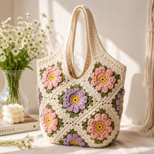 Puede incluir: Un bolso tote de ganchillo color crema construido con motivos cuadrados que presentan patrones de flores tridimensionales en tonos rosa, lavanda y amarillo con hojas verdes, acabado con dos asas integradas y un diseño de punto abierto.
