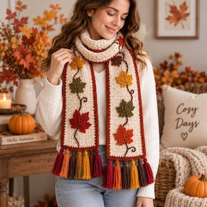 May include: A cream-colored crochet scarf featuring a dark brown vine pattern with textured, three-dimensional maple leaves in shades of yellow, olive green, and deep red. The scarf ends are finished with a row of multi-colored yarn tassels.