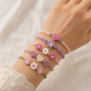 Può includere: Una collezione di braccialetti all'uncinetto in vari colori, tra cui rosa, viola e verde. Ogni braccialetto presenta un abbellimento a forma di fiore o cuore. I braccialetti sono esposti su un polso.