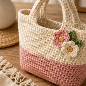 May include: A cream and pink crochet tote bag with two handles. The bag is decorated with two crochet flowers in pink and cream with green leaves. The bag is on a wooden surface. The bag is a charming handmade item.
