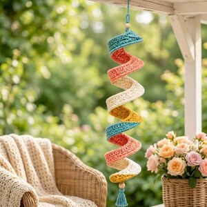 Puede incluir: Un móvil de viento en espiral hecho a mano con ganchillo. La decoración presenta bandas alternas en turquesa, coral, crema, amarillo y turquesa. Cuelga de una viga de madera con una borla turquesa. El fondo incluye una silla de mimbre y una cesta de flores.