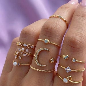 May include: A collection of gold-toned rings with various designs, including a crescent moon, stars, hearts, and floral accents. Some rings feature small gemstones. The rings are displayed on a hand against a soft purple background.