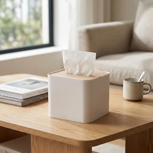 Può includere: Un dispenser di fazzoletti quadrato, beige chiaro, con coperchio in legno naturale. I fazzoletti bianchi sporgono dalla parte superiore. Il dispenser è su un tavolino di legno. Una tazza e dei libri sono visibili sullo sfondo. Design minimalista e moderno.