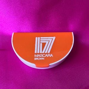 May include: A semi-circular orange and white compact case for mascara. The top of the orange lid features the white text 17, with the words MASCARA and BROWN printed in white underneath. The base of the container is white with a small orange tab.