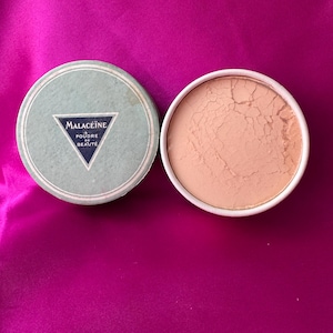May include: A round, light green container lid featuring a dark blue triangle with the text Malacéïne Poudre de Beauté, positioned next to an open circular container filled with a light beige, cracked face powder in a white base.