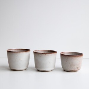 May include: Three small, handmade ceramic cups. The cups are a speckled off-white colour with a brown rim. The cups are cylindrical in shape and sit on a white surface. The cups are likely for drinking.