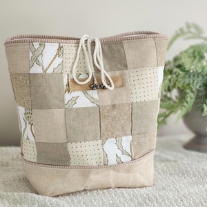 May include: A patchwork fabric bag featuring a grid of beige, tan, and white squares with botanical prints and dotted patterns. The bag has a solid light beige base, a top drawstring closure with white rope, and a small leather-like accent tab.