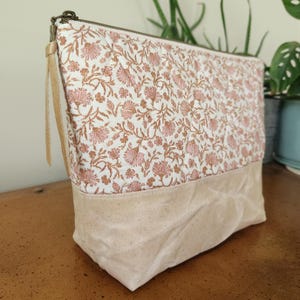 May include: A rectangular cosmetic bag with a floral pattern in pink and brown on a white background. The bottom section is a solid beige color. It has a zipper closure and a beige handle.