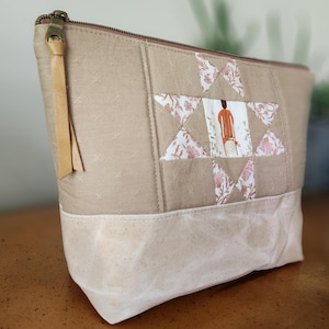 May include: Beige quilted cosmetic bag with a star-shaped patchwork design. The star features a figure in orange and white fabric. The bag has a brown zipper with a leather pull and a light beige bottom.