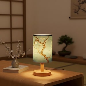 May include: A cylindrical table lamp with a light green shade featuring a tree branch design with white flowers. The lamp has a wooden base and stem. The lamp is on a wooden table.