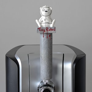 May include: A beer tap handle with a gray, textured column and a white bear figure on top. The bear has stitched details and a stitched face. The text "Tiny Rebel" is on the handle. Red drips are painted on the column.