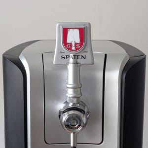 Könnte beinhalten: Ein silberner und schwarzer Bierhahn mit dem Spaten-Logo. Der Zapfhahn hat ein rot-weißes Wappen mit der Aufschrift "SPATEN München" und den Jahren "Seit 1397". Der Hahn ist Teil eines größeren Bierzapfsystems.