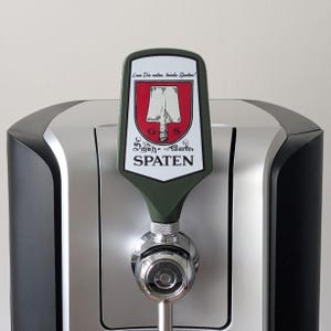 Könnte beinhalten: Ein Bierhahn mit einem grünen und weißen Spaten-Logo. Das Logo zeigt ein rotes Schild mit einer weißen Illustration und Text. Der Hahn ist an einem silbernen und schwarzen Bierausgabegerät befestigt.