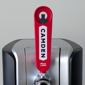May include: A red beer tap handle with the word "CAMDEN" in white, and a black castle silhouette. The handle also says "HELLS LAGER". The tap is attached to a stainless steel and black beer dispenser.