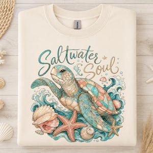 Könnte beinhalten: Ein cremefarbenes Sweatshirt mit Rundhalsausschnitt und einer grafischen Illustration einer Meeresschildkröte, die zwischen blauen Wellen, Muscheln und einem Seestern schwimmt. Der Text Saltwater Soul steht in Schreibschrift in Türkis und Gold darüber.