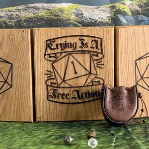 May include: Three wooden panels with engraved D&D dice shapes and the text "Crying Is A Free Action". A brown leather dice bag with the text "Best DM Dad Ever" is also present. Dice are scattered in front.