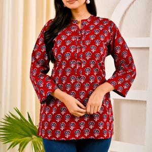 Könnte beinhalten: Eine rote Tunika mit einem Blumenmuster in Weiß und Blau. Die Tunika hat einen Stehkragen und Dreiviertelärmel. Das Model trägt eine blaue Jeans.