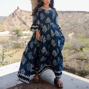 May include: A long, flowing navy blue dress with a white leaf pattern. The dress features long sleeves with flared, black and white patterned cuffs and a matching hemline. The dress is worn with brown sandals.