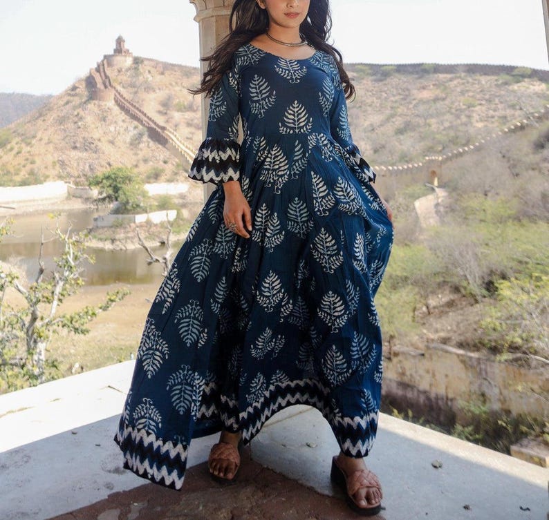 May include: A long, flowing navy blue dress with a white leaf pattern. The dress features long sleeves with flared, black and white patterned cuffs and a matching hemline. The dress is worn with brown sandals.
