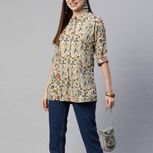 Puede incluir: Una túnica beige con botones y un estampado floral en azul, rojo y amarillo. La túnica tiene cuello mandarín y mangas tres cuartos. La modelo lleva vaqueros azul oscuro y un pequeño bolso con cuentas.