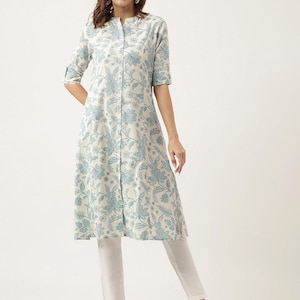 Puede incluir: Una túnica de color claro con un estampado floral en tonos azules. La túnica tiene cuello mandarín, botones frontales y mangas tres cuartos. La prenda se combina con pantalones blancos, creando un conjunto coordinado.