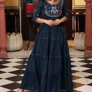 May include: A dark teal maxi dress with three-quarter sleeves and a tiered skirt. The dress features silver embellishments and intricate embroidery around the neckline. The dress is floor-length and has a flowing silhouette.