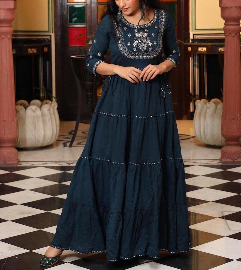 May include: A dark teal maxi dress with three-quarter sleeves and a tiered skirt. The dress features silver embellishments and intricate embroidery around the neckline. The dress is floor-length and has a flowing silhouette.
