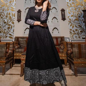 May include: A black long-sleeve dress with a flared skirt and a decorative hemline. The sleeves feature a white and black patterned design. The dress is accessorized with a silver necklace. The dress is suitable for casual or formal occasions.