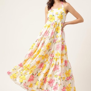 Op de afbeelding: Een witte maxi-jurk met halternek en een rok met lagen, versierd met gele en roze bloempatronen. De jurk heeft een vloeiend silhouet en een delicate rand langs de zoom, perfect voor een zomerdag.