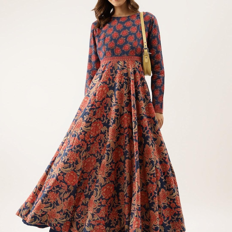 Puede incluir: Un vestido de manga larga con un estampado floral en rojo y beige sobre un fondo azul marino. El vestido tiene un cuello redondo y una cintura entallada. La falda se ensancha, creando una silueta fluida. Un peque&ntilde;o bolso de color claro cuelga del hombro.