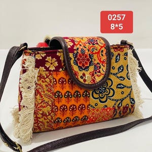 Könnte beinhalten: Bunte Umhängetasche mit Patchwork-Design. Die Tasche hat einen Überschlag und einen braunen Lederriemen. Die Tasche misst 20 cm x 13 cm und hat Fransen an den Seiten. Die Tasche hat ein Muster aus Blumen und geometrischen Formen.