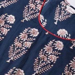 May include: Close-up of a navy blue garment with a floral pattern in white and burgundy. The design includes large flower and leaf motifs with sequin accents. A red trim outlines the neckline.