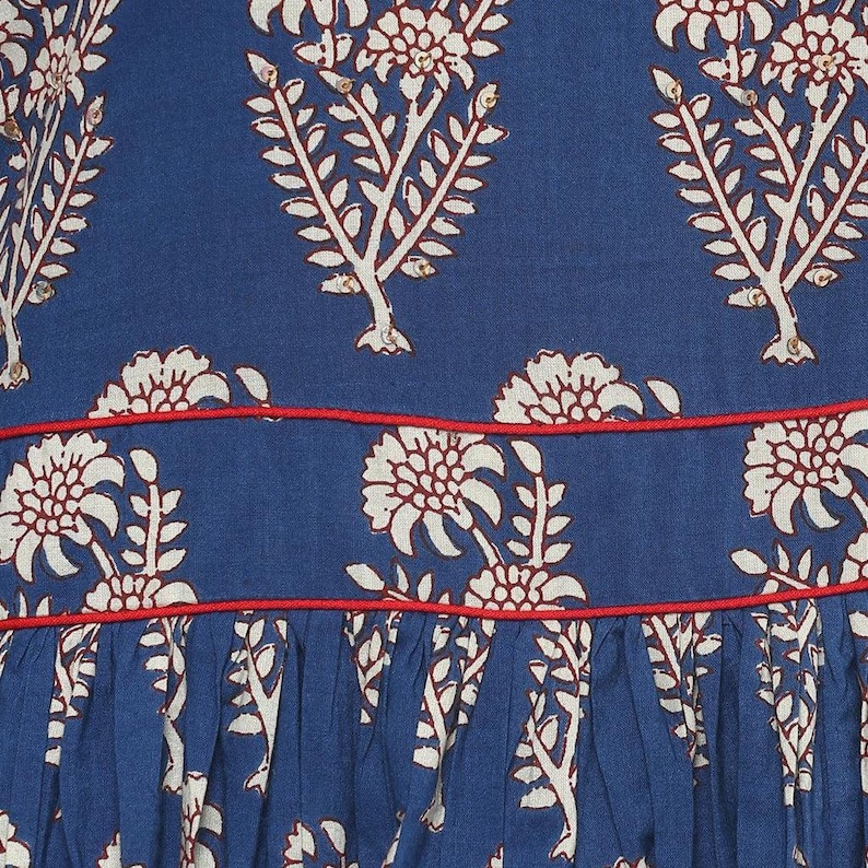 May include: Close-up of a blue fabric with a repeating floral pattern in white and red. The design features stylized flowers and leafy branches. A thin red line accents the fabric's design.