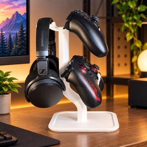 Könnte beinhalten: Ein weißer vertikaler Ständer mit quadratischem Fuß hält einen schwarzen Sennheiser-Kopfhörer auf der linken Seite, einen einfarbig schwarzen Gamecontroller oben und einen schwarzen Gamecontroller mit roten Flammengrafiken unten rechts.