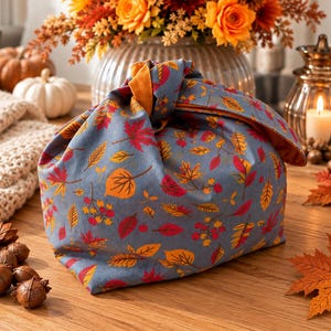 Könnte beinhalten: Eine Bento-Tasche aus Stoff mit blauem Hintergrund und einem wiederkehrenden Muster aus Herbstblättern in Rot-, Orange- und Gelbtönen. Die Tasche ist oben verknotet, wobei das einfarbige orangefarbene Innenfutter an der Öffnung sichtbar ist.
