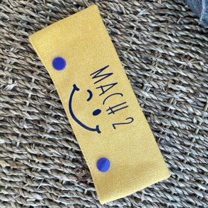 Könnte beinhalten: Gelbe rechteckige Tasche mit dem Text "MACH 2" und einem marineblauen Smiley-Gesicht. Zwei marineblaue runde Knöpfe sichern die Tasche. Die Tasche liegt auf einer gewebten Oberfläche.