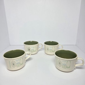 May include: Four cream ceramic mugs with olive green interiors. Each mug features a mid-century modern design with a starburst and geometric shapes in black, light blue, and green. The mugs have a curved shape and a handle.