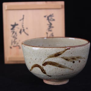 以下が含まれることがあります： 外側に手描きの濃い茶色の葉のデザインが施された、斑点のある明るいベージュの陶器製茶碗。茶碗は、蓋に黒い日本語の書道文字が書かれた正方形の木製収納ボックスの前に置かれています。