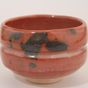 May include: A round, hand-thrown ceramic bowl with a glossy, reddish-orange glaze. The exterior features irregular, dark charcoal-black abstract markings and a horizontal indentation circling the middle. The base is a contrasting, unglazed light beige.