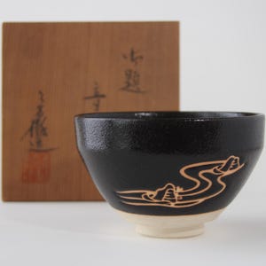 May include: A black ceramic tea bowl with a textured surface and a light-colored base. The bowl features a gold-colored line drawing of a winding river and mountains. Behind the bowl sits a square wooden box with black Japanese calligraphy.