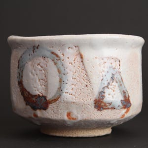 May include: A ceramic tea bowl with a textured, off-white glaze. The exterior features hand-painted geometric shapes, including a circle and a triangle, in muted blue and brown tones. The surface has a porous, speckled appearance with drips.