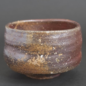 May include: A small, round ceramic bowl with a thick, uneven rim. The exterior features a textured, mottled surface with deep brown, reddish-brown, and earthy tan tones. The glaze appears weathered with visible speckles and subtle horizontal ridges.