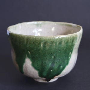 May include: A ceramic bowl with a speckled off-white base and a thick, uneven forest green glaze dripping from the rim down the sides. The bowl features a rounded, organic shape with a textured surface and a small, circular footed base.