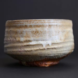 May include: A ceramic tea bowl with a cylindrical shape and a textured, uneven surface. The exterior features a mottled glaze in shades of cream, beige, and light brown, with a darker, reddish-brown base and visible horizontal ridges along the side.