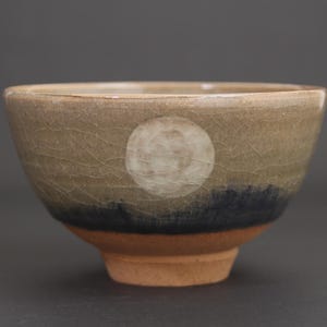 May include: A ceramic bowl with a crackle glaze finish featuring a beige upper section, a dark blue band, and a natural terracotta base. A circular white moon design is centered on the beige portion of the bowl, creating a landscape effect.