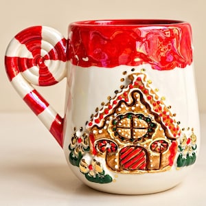 Könnte beinhalten: Ein Keramikbecher mit cremefarbener Basis und einem erhabenen Lebkuchenhaus-Design auf der Vorderseite. Der Henkel ist wie ein rot-weiß gestreiftes Pfefferminzbonbon geformt. Der Rand ist mit einer glänzenden, strukturierten roten Glasur bemalt.