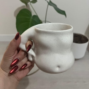 Puede incluir: Una taza de cerámica blanca con acabado moteado y forma orgánica redondeada. La taza presenta un asa curva prominente y una pequeña hendidura en relieve en la base, que recuerda a un torso humano con un ombligo y contorno de cadera.