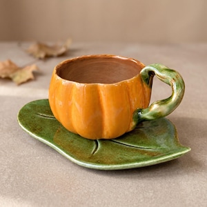 Könnte beinhalten: Eine Keramiktasse in Form eines leuchtend orangefarbenen Kürbisses mit glänzender Oberfläche und einem gedrehten grünen Henkel, die auf einer passenden grünen Untertasse in Blattform mit sichtbaren Blattadern steht. Das Innere ist naturfarben.