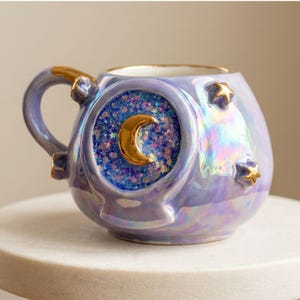 May include: A round ceramic mug with an iridescent lavender finish. The mug features a central circular inset filled with blue glitter and a raised gold crescent moon. Additional gold star accents are applied to the mug body and handle.