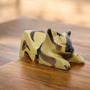 May include: A ceramic cat figurine with a glossy finish, featuring a light tan base color with dark brown, abstract brushstroke markings. The cat is depicted in a resting, prone position with its head slightly raised and ears pointed forward.